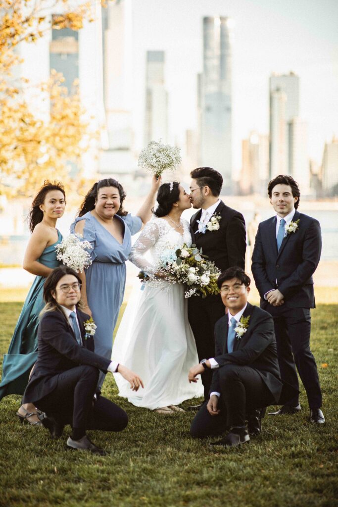 Bride and groom with their wedding party posing on the lawn with the city skyline in the background. NJ Micro Wedding Photographer for intimate weddings, courthouse weddings, backyard weddings, and elopements.
