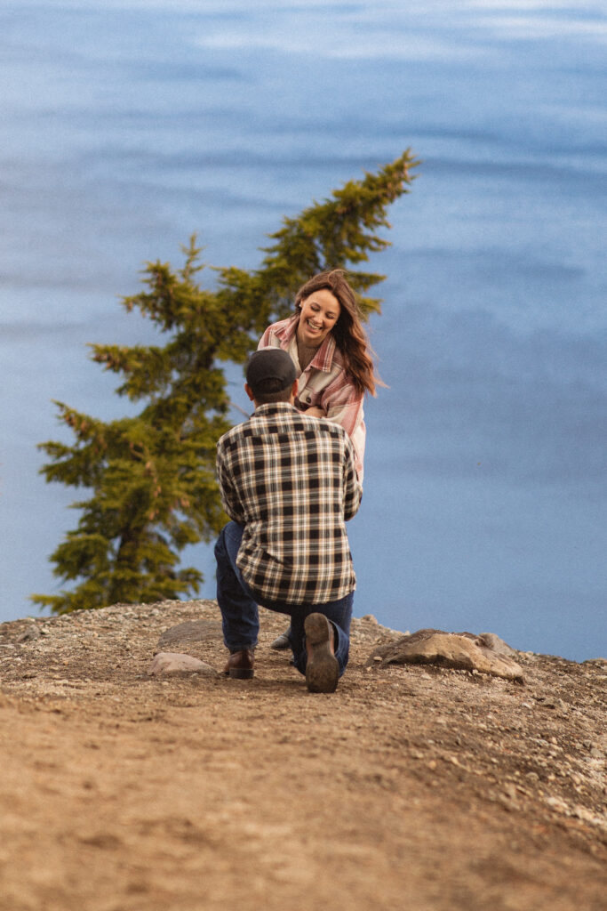 Surprise Reaction: Cassie smiling and covering her face in joy as Ronnie proposes to her at Crater Lake.

