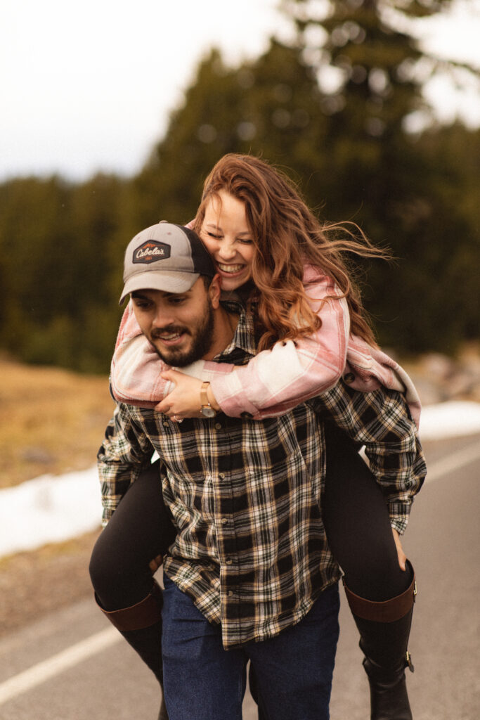 Another Piggyback Ride: Cassie laughing joyfully on Ronnie’s back as they walk down a forest road, celebrating their engagement.

