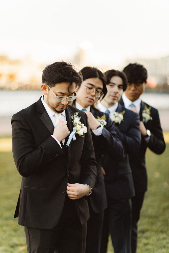 Groomsmen standing in line with the groom, each adjusting their boutonniere. New Jersey documentary wedding photographer capturing candid moments.
