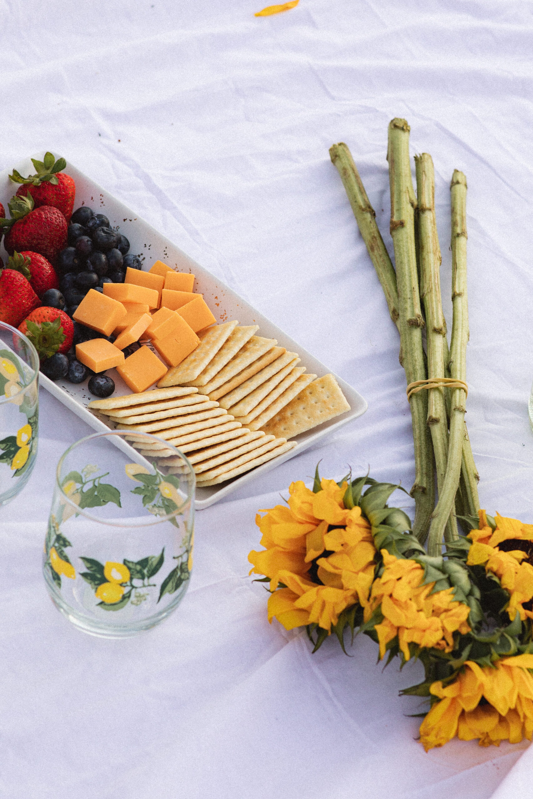 Close-up of a picnic spread featuring crackers, cheese, strawberries, and sunflowers on a white blanket.