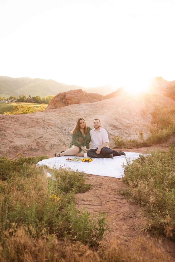 The couple sits on a white blanket, enjoying their picnic with the sun setting in the background, creating a peaceful scene.