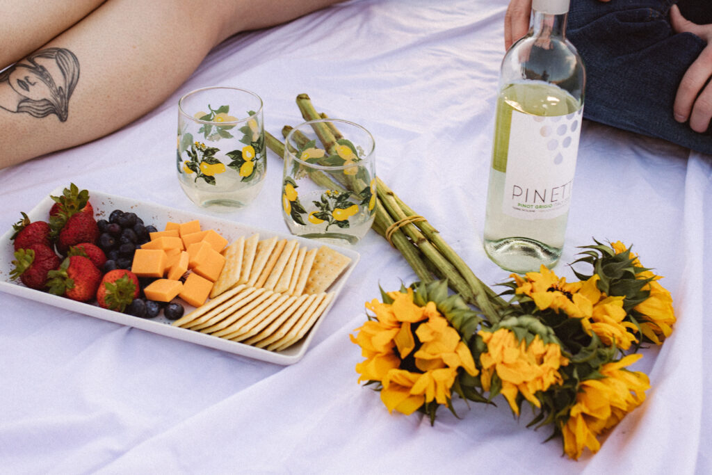 A close-up of the picnic setup, with a white sheet, cheese, crackers, sunflowers, and two decorative glasses.