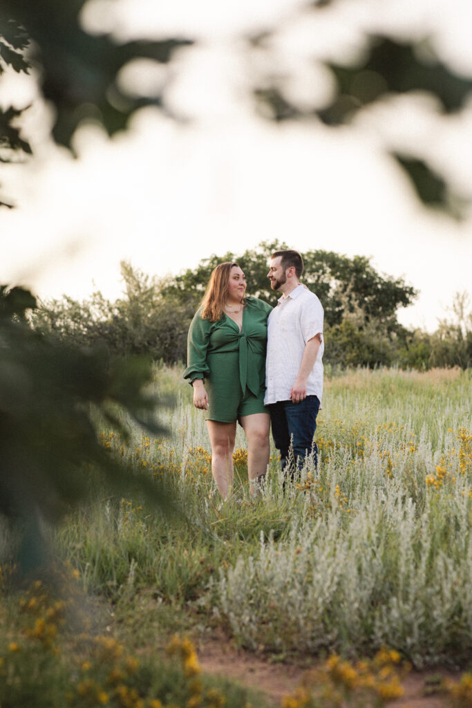 The couple stands close together in a grassy field, framed by foliage, capturing a serene moment.