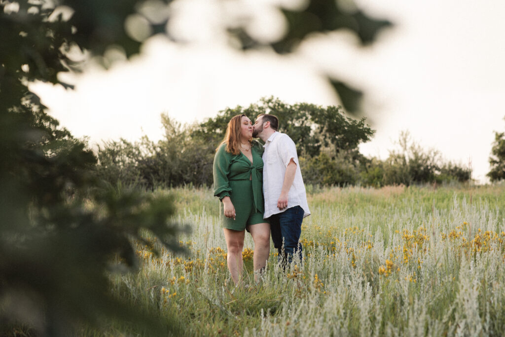 The couple kisses in a grassy field, with the sun setting behind them, creating a romantic atmosphere.