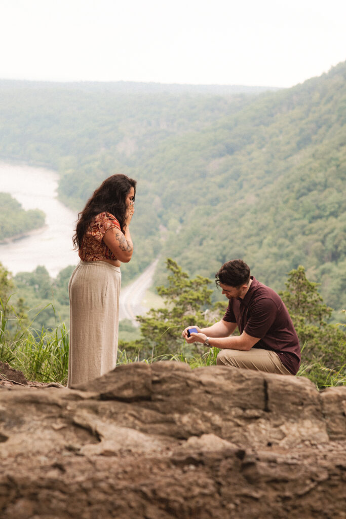 Alec proposes to Jac on a scenic overlook at Mount Tammany, surrounded by lush greenery and the Delaware Water Gap.

