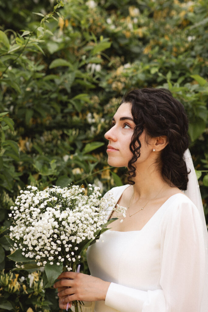 Bride holding a bouquet of white flowers, standing in front of green foliage.