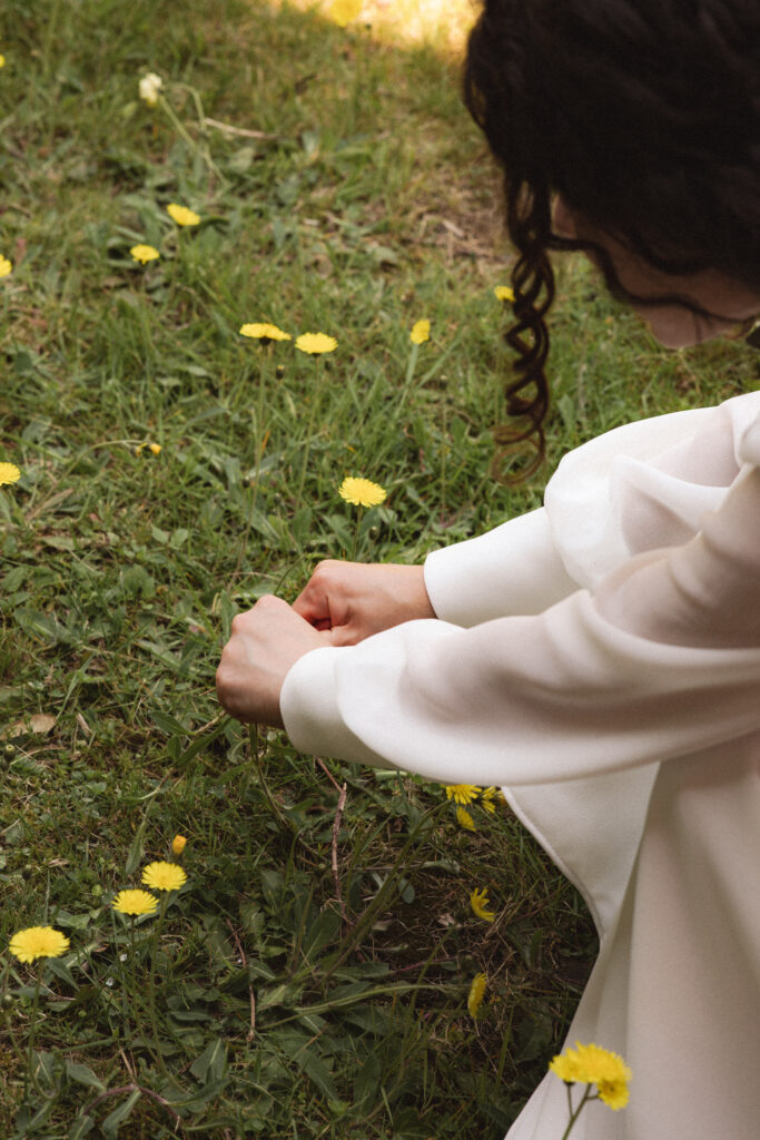 Bride sitting on the grass, adjusting her dress, holding a flower.
