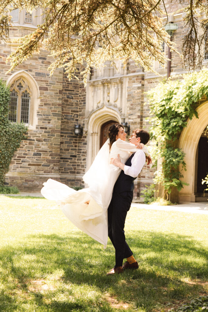 Groom lifting the bride in front of a cathedral, both smiling joyfully.
