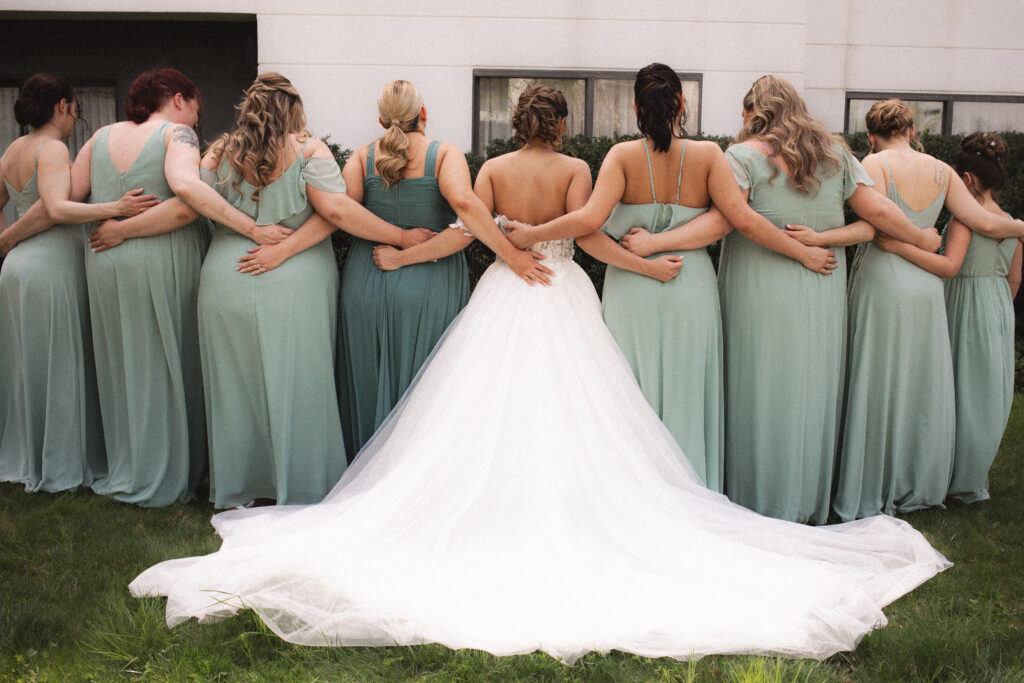 Bride Briana with her bridesmaids, all showing the backs of their dresses, standing on grass.