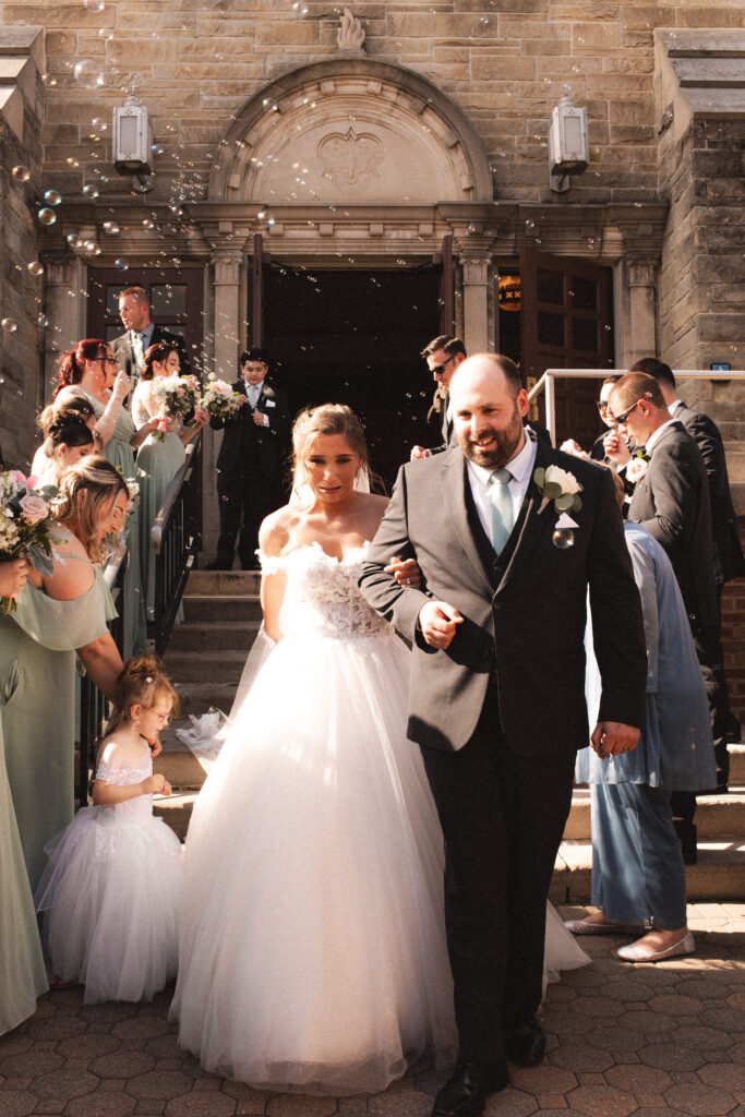 Briana and Pat smiling and walking arm in arm outside the church, surrounded by guests.