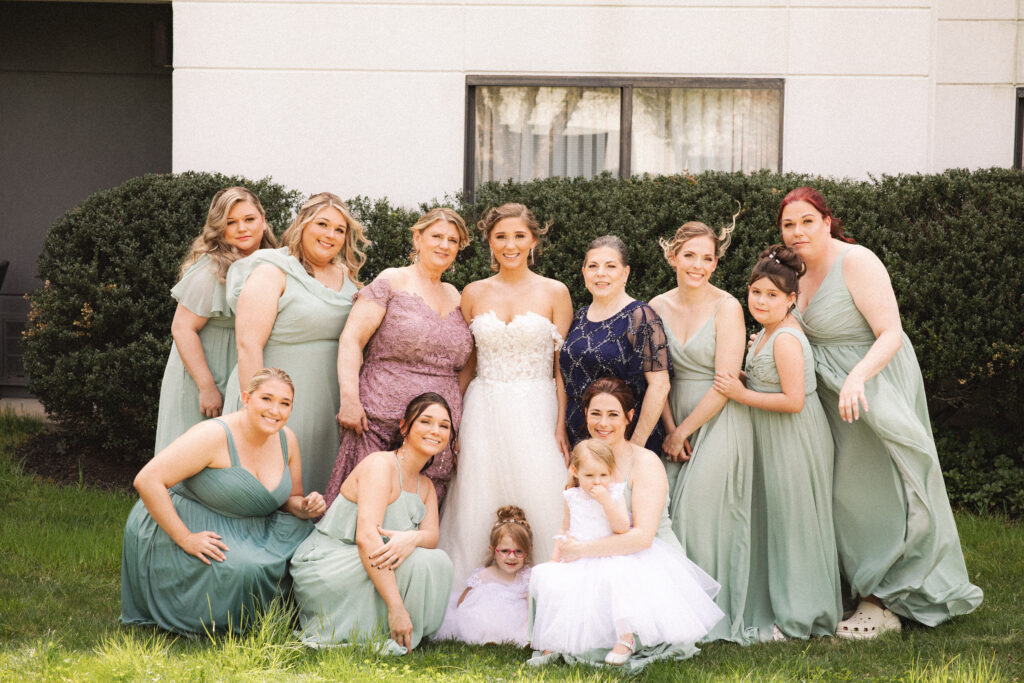 Briana posing with her bridesmaids, all in light green dresses, outside on a grassy area.

