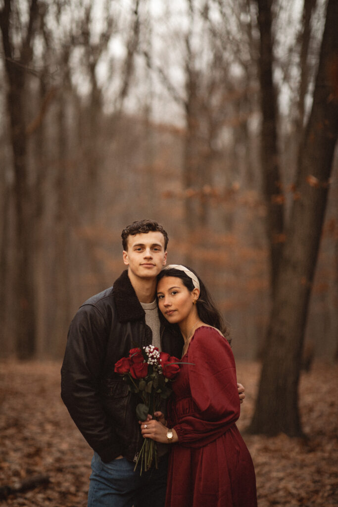 Couple embracing with gentle smiles, the woman holding a bouquet of roses. Scenic background in the forest. Intimate photography session