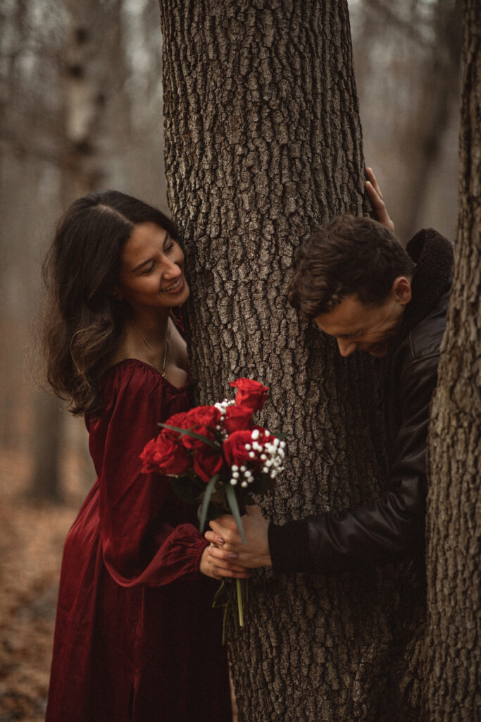 Couple exchanging smiles while leaning against a tree, with the woman holding roses.
