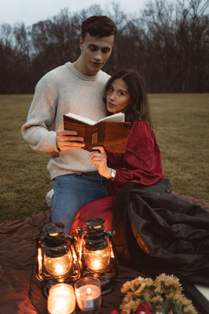 Couple sitting on a blanket in a field, reading a book together by lantern light.